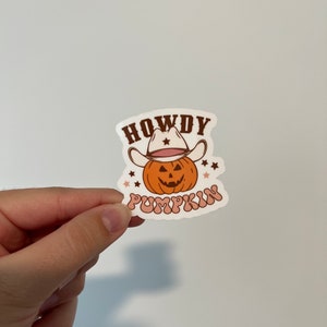 May include: A sticker featuring a cartoon pumpkin wearing a cowboy hat. The pumpkin is orange with a carved face. The text "HOWDY PUMPKIN" is written in brown and pink. Small stars are scattered around the pumpkin.