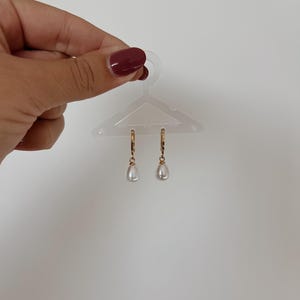 May include: Gold-tone hoop earrings with teardrop-shaped faux pearls. The earrings are displayed on a clear plastic hanger against a white background. The earrings are a classic jewelry piece.