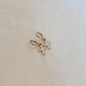 May include: A pair of gold-colored hoop earrings with white cross pendants. The earrings are displayed on a light-colored surface. The crosses have a white enamel fill and a gold outline.