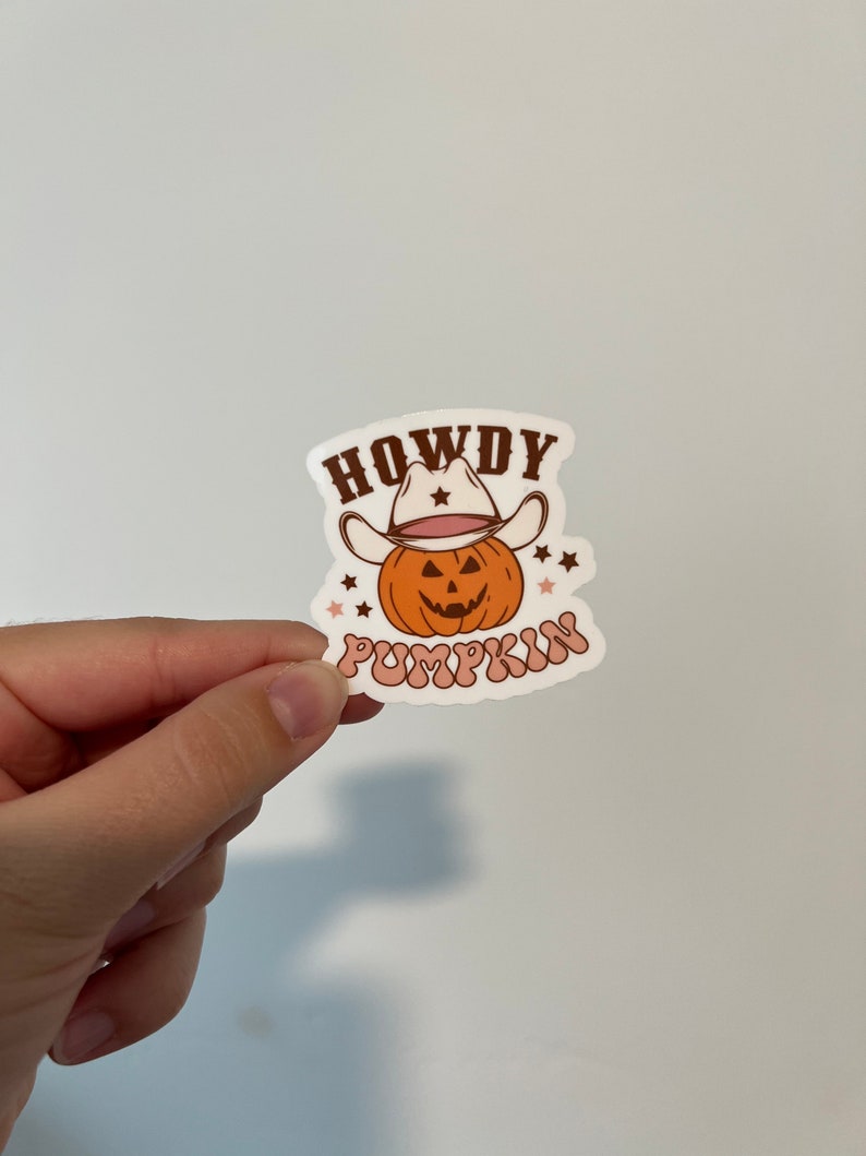 May include: A sticker featuring a cartoon pumpkin wearing a cowboy hat. The pumpkin is orange with a carved face. The text "HOWDY PUMPKIN" is written in brown and pink. Small stars are scattered around the pumpkin.