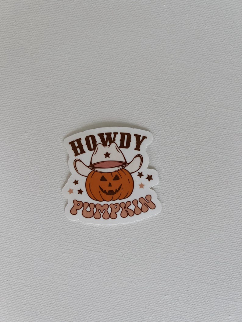 May include: A sticker featuring a cartoon pumpkin wearing a cowboy hat. The pumpkin is orange with a carved face. The text "HOWDY PUMPKIN" is written above and below the pumpkin in a brown, retro font. Small stars surround the pumpkin.