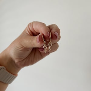 May include: A pair of gold hoop earrings with a small key charm hanging from each hoop. The keys have a heart-shaped keyhole.