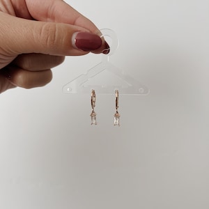 May include: Rose gold hoop earrings with rectangular, clear crystal pendants. The earrings are displayed on a clear plastic hanger against a white background. The earrings are held by a hand with a maroon nail polish.