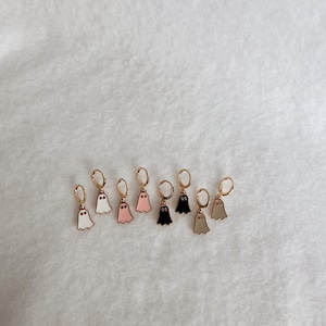 May include: A collection of gold-colored hoop earrings, each featuring a small, ghost-shaped charm in various colors: white, pink, black, and beige. The earrings are displayed on a white, textured surface.