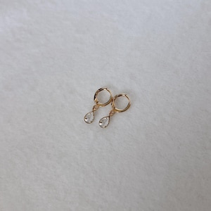 May include: A pair of gold hoop earrings featuring small, clear, teardrop-shaped charms suspended from each hoop. The earrings have a polished metallic finish and a simple, delicate design, with the clear stones catching the light.