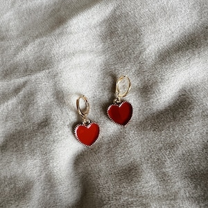 May include: A pair of gold-colored hoop earrings with red heart-shaped pendants. The heart pendants have a red enamel finish with a gold-colored border. The earrings are displayed on a gray, textured surface.