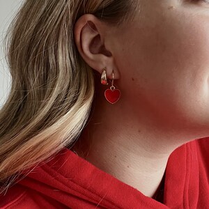 May include: A pair of gold hoop earrings and a gold hoop earring with a red heart charm. The earrings are worn on the ear of a person wearing a red hoodie.
