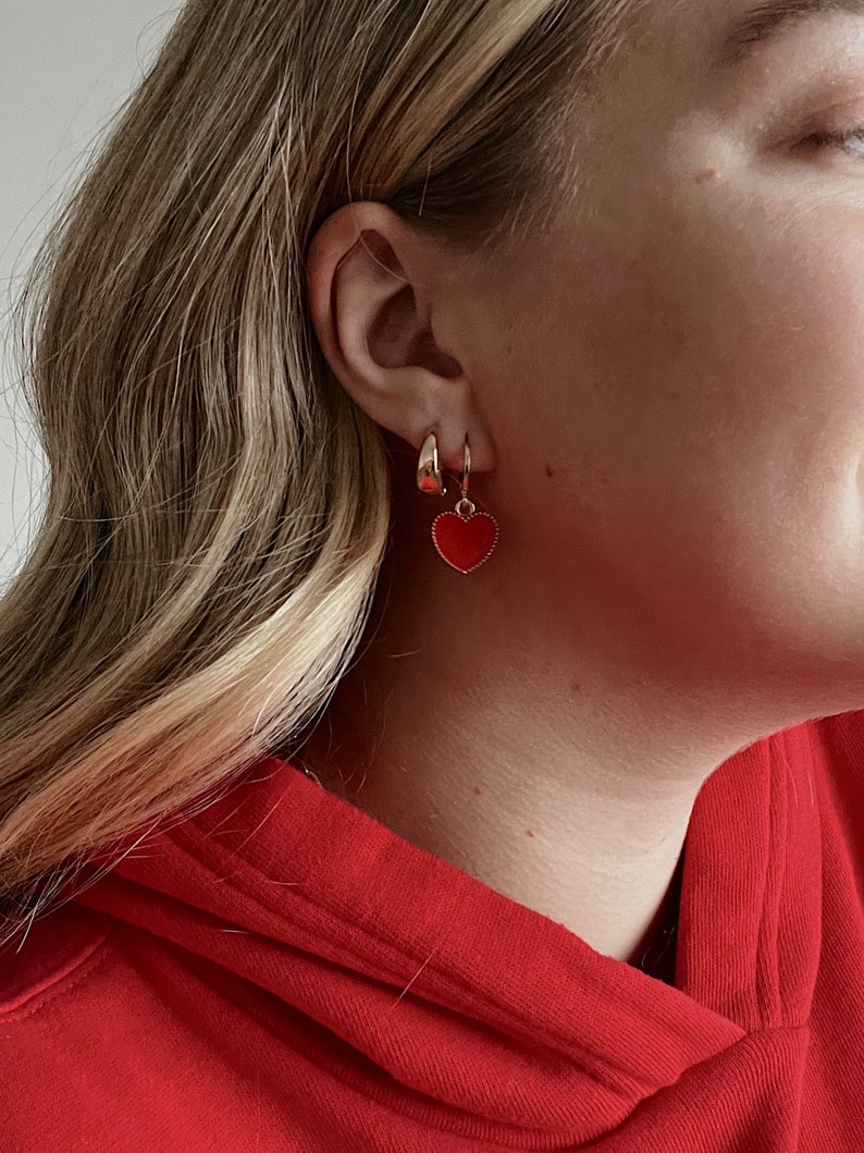 May include: A pair of gold hoop earrings and a gold hoop earring with a red heart charm. The earrings are worn on the ear of a person wearing a red hoodie.