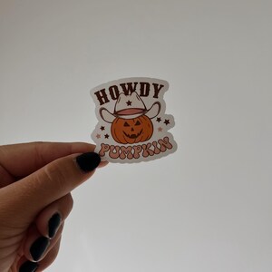 May include: A sticker featuring a smiling pumpkin wearing a cowboy hat. The text "HOWDY PUMPKIN" is arched above and below the pumpkin. The design has a retro, vintage aesthetic with orange, brown, and cream colors.