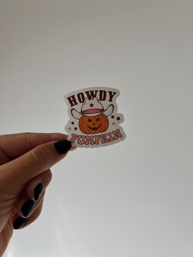 May include: A sticker featuring a smiling pumpkin wearing a cowboy hat. The text "HOWDY PUMPKIN" is arched above and below the pumpkin. The design has a retro, vintage aesthetic with orange, brown, and cream colors.