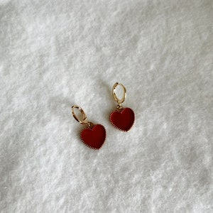 May include: A pair of gold-colored hoop earrings with red heart-shaped pendants. The heart pendants have a textured border and are filled with a solid red color. The earrings are displayed on a white, textured surface.
