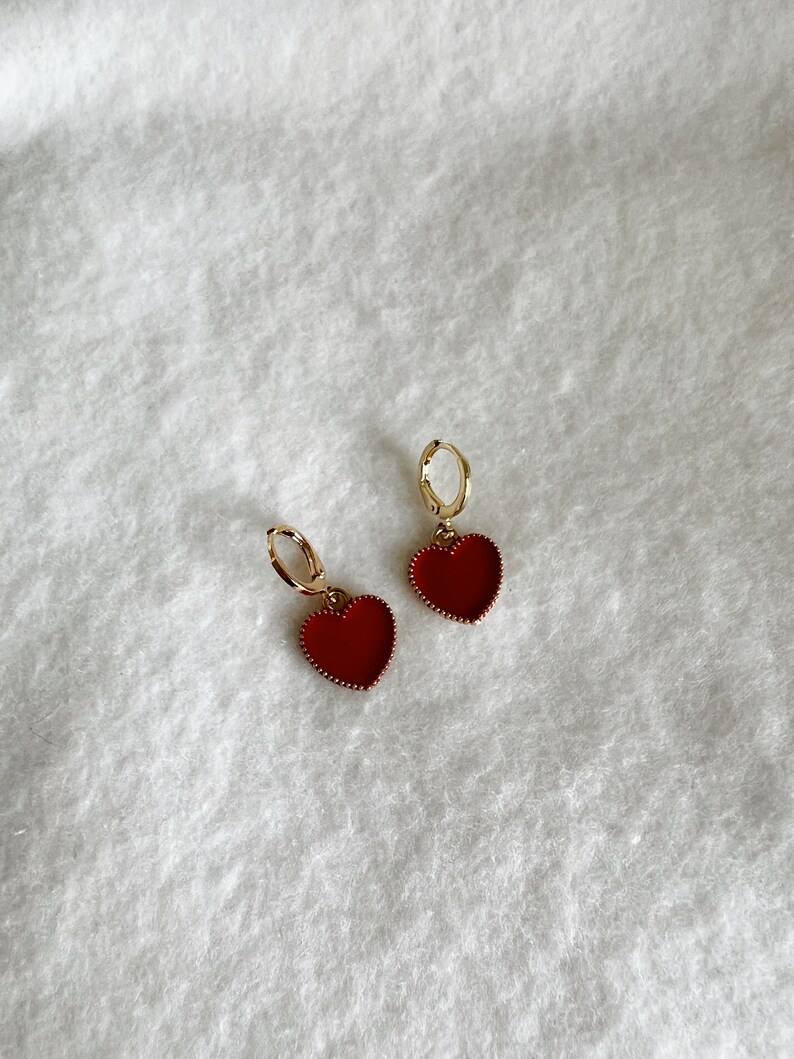 May include: A pair of gold-colored hoop earrings with red heart-shaped pendants. The heart pendants have a textured border and are filled with a solid red color. The earrings are displayed on a white, textured surface.