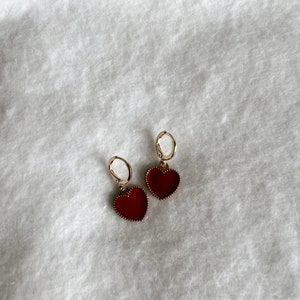 May include: A pair of gold-colored hoop earrings with heart-shaped red pendants. The earrings are displayed on a white, textured surface. The heart pendants have a detailed border.