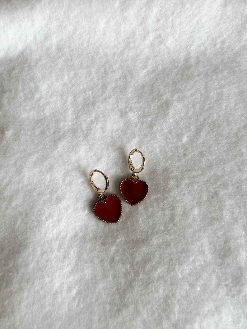 May include: A pair of gold-colored hoop earrings with heart-shaped red pendants. The earrings are displayed on a white, textured surface. The heart pendants have a detailed border.