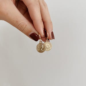 May include: A pair of gold hoop earrings with a coin charm. The coin has a profile of a woman on one side and a design with a crown on the other side.