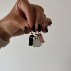 May include: Three ghost-shaped earrings in black, white, and pink, each with a gold-colored hoop. The earrings are held up against a plain white background. The ghost earrings are a fun accessory.