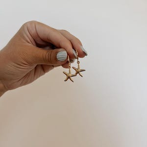 May include: Gold-colored starfish earrings. Each earring features a small, detailed starfish charm suspended from a hoop. The earrings are held against a neutral background, highlighting their design and color.
