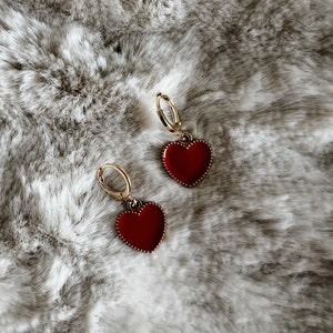 May include: A pair of gold hoop earrings with red heart-shaped pendants. The heart pendants have a glossy finish and a beaded border. The earrings are displayed on a soft, textured, gray and white surface.
