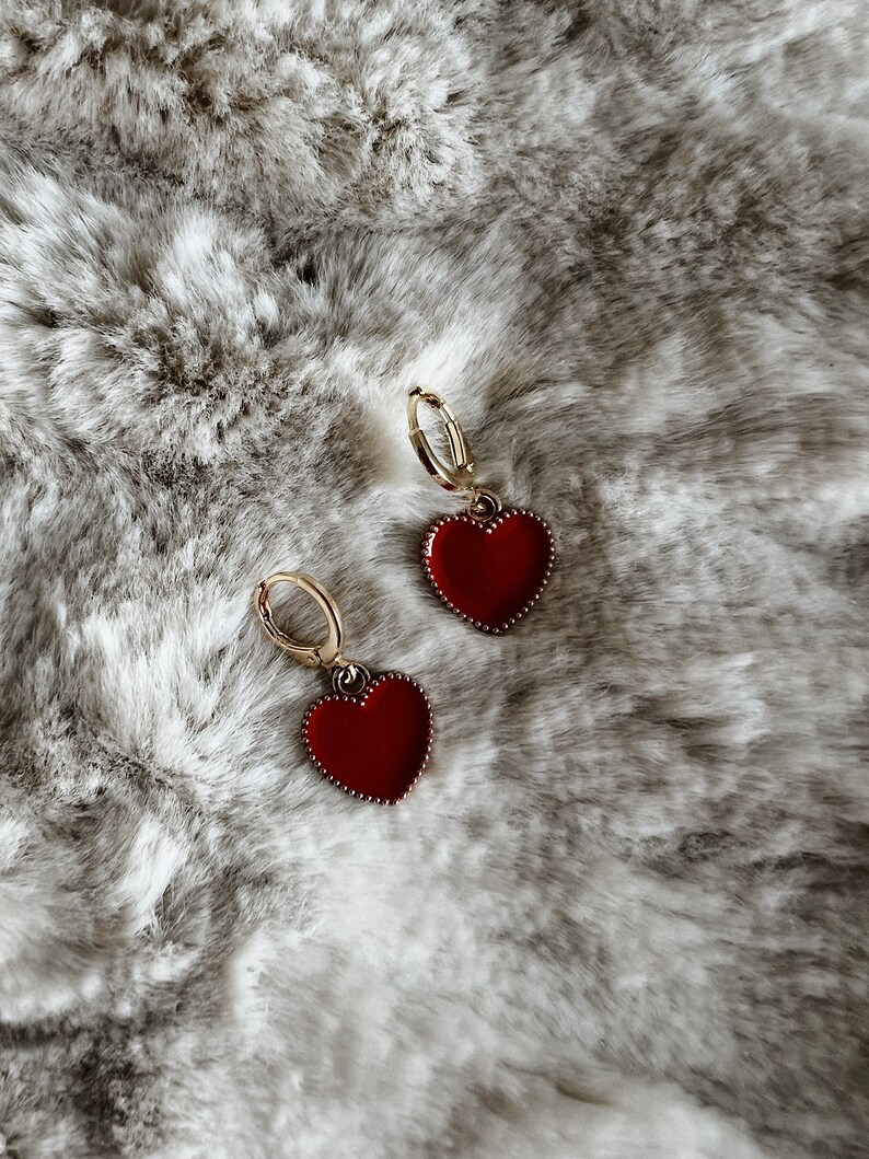 May include: A pair of gold hoop earrings with red heart-shaped pendants. The heart pendants have a glossy finish and a beaded border. The earrings are displayed on a soft, textured, gray and white surface.
