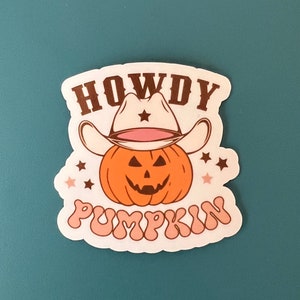 May include: A sticker featuring a cartoon pumpkin wearing a cowboy hat. The pumpkin is orange with a carved face. The text "HOWDY PUMPKIN" is written above and below the pumpkin in brown and pink.
