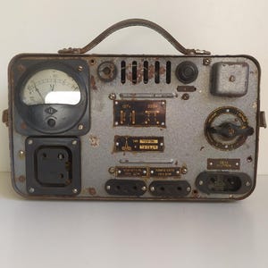 Può includere: Dispositivo elettrico vintage in metallo grigio con maniglia, indicatori, quadranti e prese multiple. Il dispositivo ha un'estetica industriale usurata, con evidenti segni di età e utilizzo. Include un indicatore circolare, un interruttore rotante e vari punti di connessione.