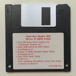 Puede incluir: Un disquete negro de 3,5 pulgadas con una cubierta metálica plateada y una etiqueta blanca. La etiqueta presenta texto en rojo que dice Intel Bus Master IDE Driver & BIOS Utility, enumerando la versión 3.01, utilidades y el número de pieza P/PII-IDE-V5.0.