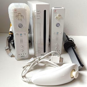 Peut inclure: Une console Nintendo Wii blanche avec deux télécommandes blanches, un nunchuk et une barre de capteurs. La console a un design vertical avec un bouton d'alimentation et un bouton de réinitialisation. Les manettes ont un pavé directionnel en forme de croix et plusieurs boutons.