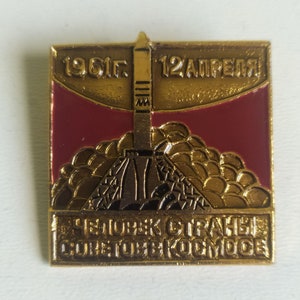 May include: A gold-colored pin with a red background. The pin depicts a rocket launching into the sky with the text "1961г. 12АПРЕЛЯ" and "ЧЕЛОВЕК СТАРАИС СОВЕТСКОГО КОСМОСА".