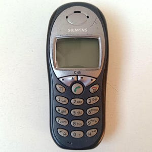 Op de afbeelding: Een zwart en zilver Siemens C45 mobiele telefoon. De telefoon heeft een klein scherm, een toetsenbord met cijfers en symbolen en het Siemens-logo bovenaan. Een klassiek ontwerp uit de vroege jaren 2000.