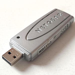 May include: Silver NETGEAR WG111v3 USB wireless adapter. The adapter has a USB connector and the NETGEAR logo is embossed on the top. The device is rectangular and has a small indicator light.