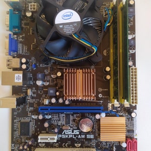 May include: A close-up of a brown and gold ASUS P5KPL-AM SE motherboard with an Intel processor, a black heatsink with a fan, and two green RAM sticks. The motherboard has a blue PCI Express slot, a black battery, and a blue IDE connector.