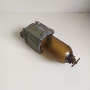 Op de afbeelding: Een vintage brandstoffilter van metaal en glas met een bruin glazen lichaam en een metalen bovenkant met een schroefdop. Het filter heeft een klein metalen handvat aan de zijkant.