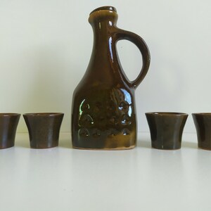 Può includere: Un set di recipienti per bere in ceramica marrone, tra cui una brocca e quattro piccole tazze. La brocca ha un manico e una superficie testurizzata.