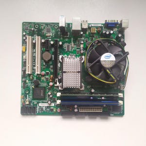 May include: A green computer motherboard with a black and silver CPU cooler. The cooler has a fan with an Intel logo. The motherboard has several slots for RAM, a network port, and a VGA port.
