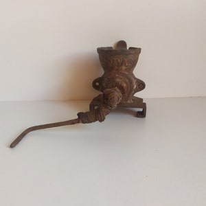 May include: A vintage cast iron meat grinder with a rusty brown finish. The grinder has a crank handle and a metal spout. The grinder is mounted on a wall.