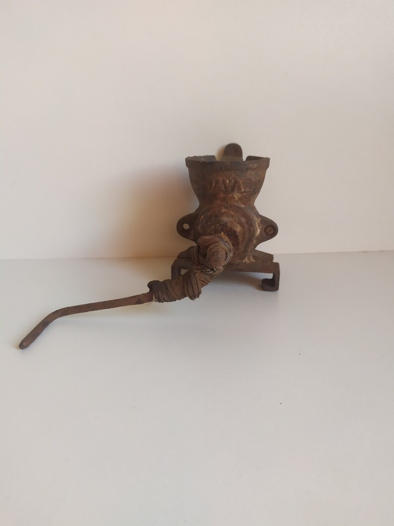 May include: A vintage cast iron meat grinder with a rusty brown finish. The grinder has a crank handle and a metal spout. The grinder is mounted on a wall.