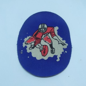 Könnte beinhalten: Ein blauer und weißer Stoff-Patch mit einem Cartoon-Bild eines Footballspielers in einem roten Trikot und einem weißen Helm, der mit dem Ball rennt. Der Patch ist oval geformt und hat einen weißen Hintergrund mit blauen Farbspritzern.
