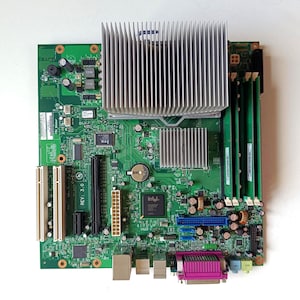 Op de afbeelding: Een groen computermoederbord met een grote zilveren aluminium koelplaat, drie groene RAM-modules, een centrale Intel-processorchip, twee witte PCI-sleuven, een paarse parallelle poort en diverse geïntegreerde elektronische componenten.