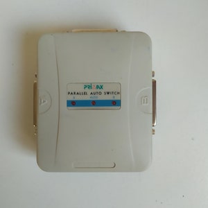 以下が含まれることがあります： 前面に「PRIMAX」と「PARALLEL AUTO SWITCH」の文字が印刷された白いパラレルオートスイッチ。スイッチには「A」と「B」のラベルが付いた2つのポートと、「AUTO」のラベルが付いた3つの赤いLEDがあります。