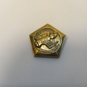 Könnte beinhalten: Eine goldfarbene Metallstifte mit einer Fünfecksform. Die Stifte zeigt ein stilisiertes Bild eines Raumschiffs mit dem Text "Космический корабль "Союз" in kyrillischer Schrift.