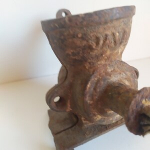 May include: A vintage cast iron coffee grinder with a crank handle. The grinder is rusty and has a worn surface. The grinder has a hopper on top and a base with a small platform.