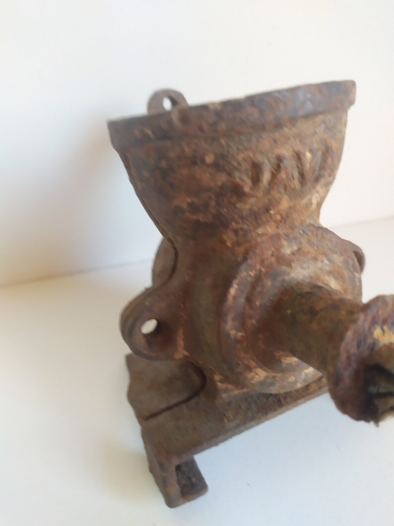 May include: A vintage cast iron coffee grinder with a crank handle. The grinder is rusty and has a worn surface. The grinder has a hopper on top and a base with a small platform.