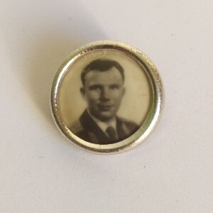 May include: A silver-toned round pin with a black and white photo of a man in a suit and tie inside.