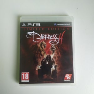 Könnte beinhalten: Ein Videospiel-Etui für The Darkness II, ein PlayStation 3-Spiel. Das Etui ist schwarz mit einem roten und weißen Design. Das Spiel ist ab 18 Jahren freigegeben und ein Titel von 2K Games.