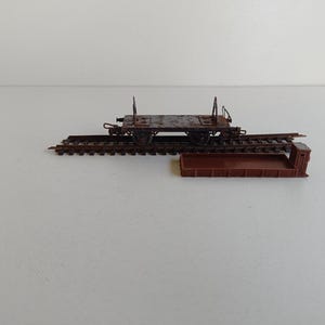 May include: A miniature model of a rusty brown railroad flatcar with a small brown platform and a section of track. The flatcar has four wheels and a flat bed. The track is made of brown metal.