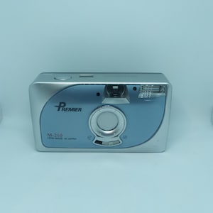 May include: A silver and blue compact film camera with the brand name "PREMIER" and the model number "M-210" printed on the front. The camera has a lens made in Japan.