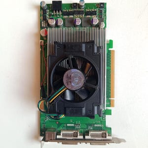 May include: A green and black computer graphics card with a black fan in the center. The card has multiple ports and connectors, and a silver heat sink. The fan has a multi-colored wire.