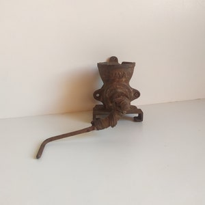 Può includere: Un macinaca carne a manovella in ghisa arrugginita montato a parete con un lungo braccio di manovella in metallo.