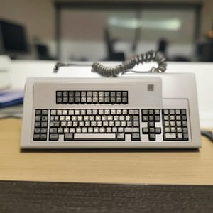 Op de afbeelding: Een vintage, lichtgrijs toetsenbord met een telefoonsnoer. Het toetsenbord heeft een rechthoekig ontwerp met meerdere toetsen en een kleiner toetsenbord boven het hoofdtoetsenbord. Het staat op een houten bureau.