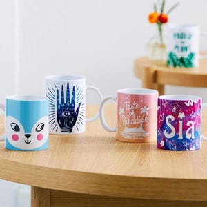 May include: Four ceramic mugs with white handles are displayed on a wooden table. One mug is light blue with a fox face, another has a blue hand design, a third has "Taste of Paradise" text, and the last has "Sia" text. A fifth mug is visible in the background.